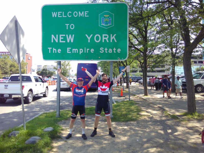 Chris and James make a Y sign with their arms in front of the Welcome to New York sign
